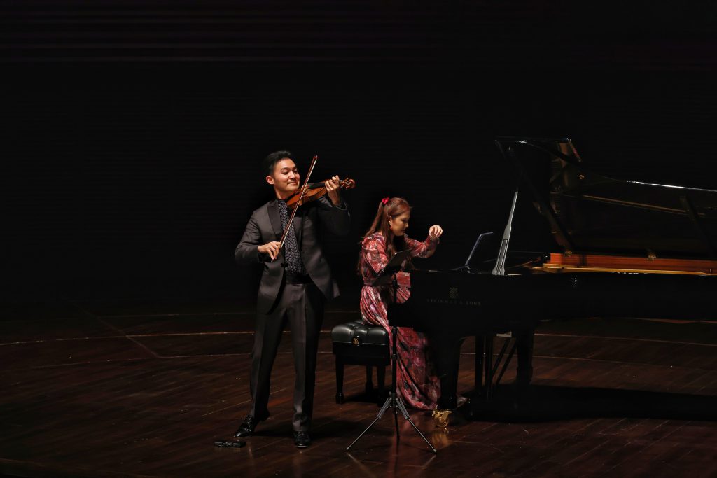 Violinist in a dark suit plays violin while a pianist in a patterned dress accompanies at a grand piano on a dimly lit stage.