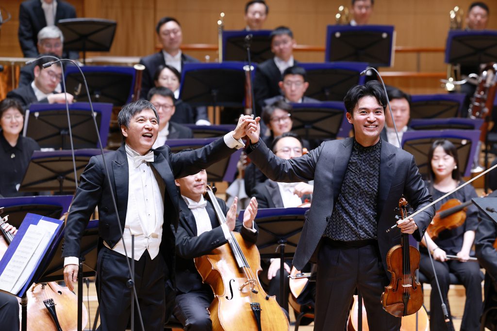 Two male cellists in tuxedos raise their hands in celebration in front of a full orchestra on stage.
