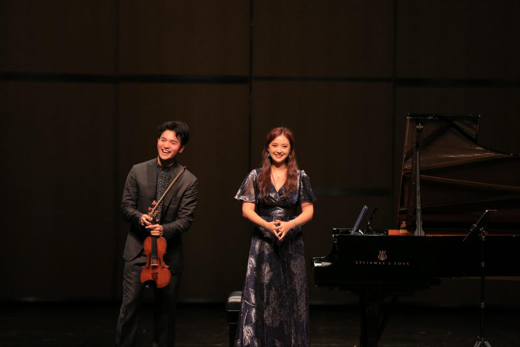 Male violinist and female vocalist stand on a concert stage beside a grand piano.