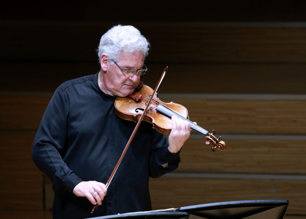 Older man with white hair playing a violin on stage, focused on the music.