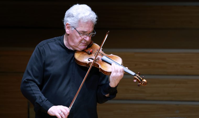 Older man with white hair playing a violin on stage, focused on the music.