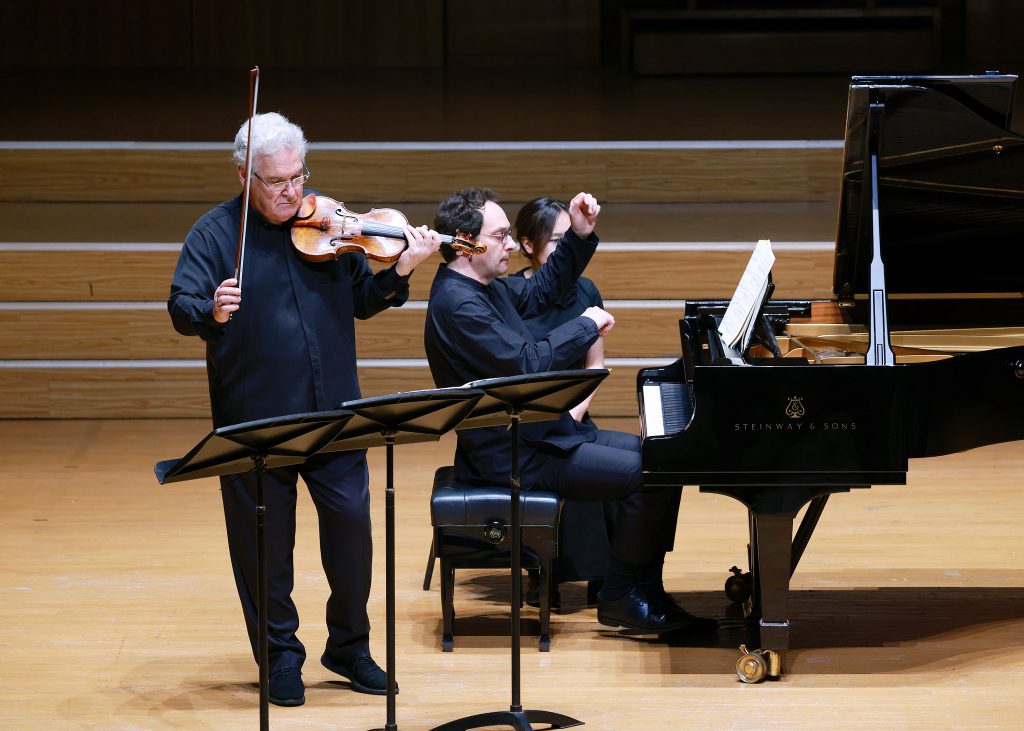 Violinist standing with bow raised while two musicians sit at a grand piano reading sheet music on a wooden stage.