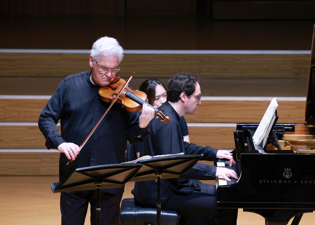 Violinist with white hair playing violin while a pianist accompanies on a grand piano on stage.
