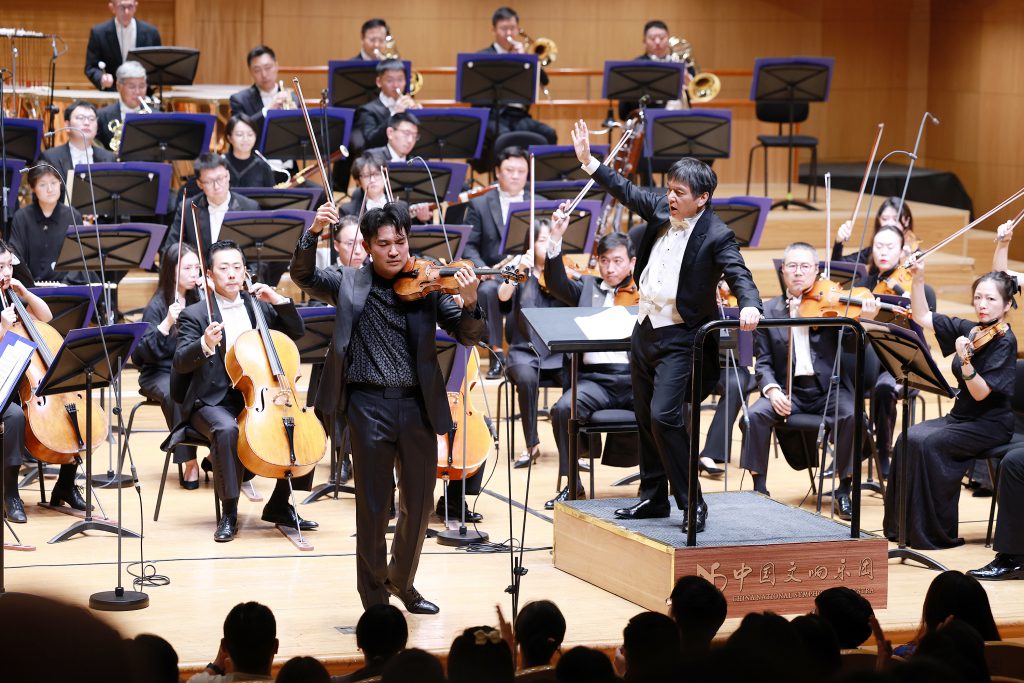 Conductor leads a full symphony orchestra on stage while a violinist performs in the foreground during a concert.