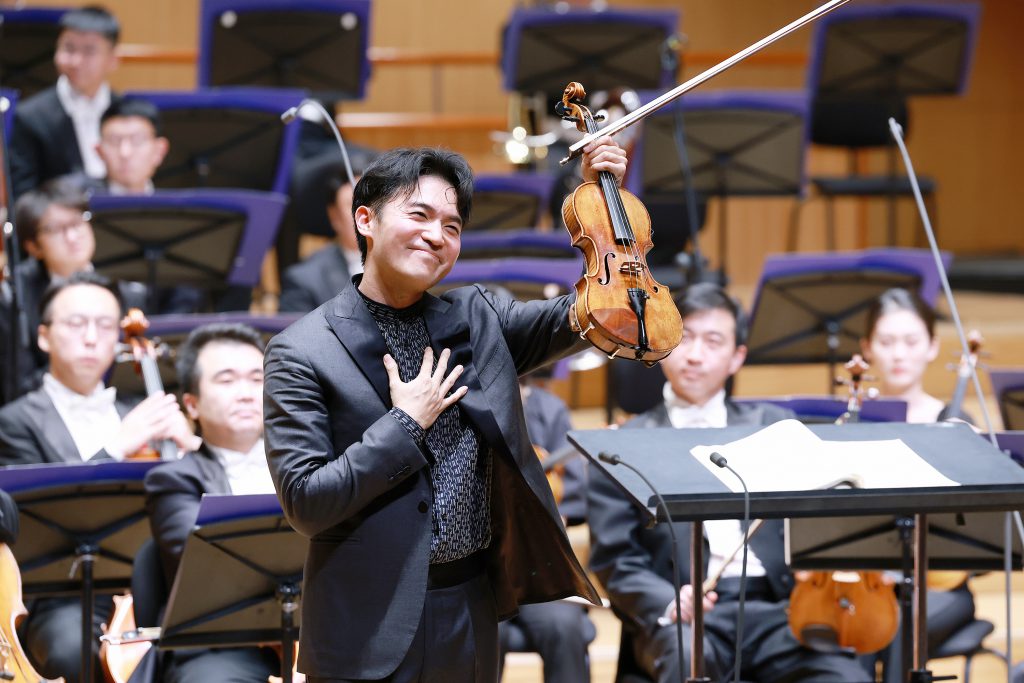 Violinist in a black suit raises his violin and bow while smiling; the orchestra sits behind him on stage.