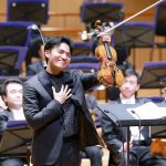 Violinist in a black suit raises his violin and bow while smiling; the orchestra sits behind him on stage.