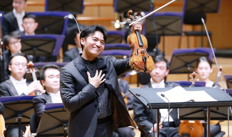 Violinist in a black suit raises his violin and bow while smiling; the orchestra sits behind him on stage.