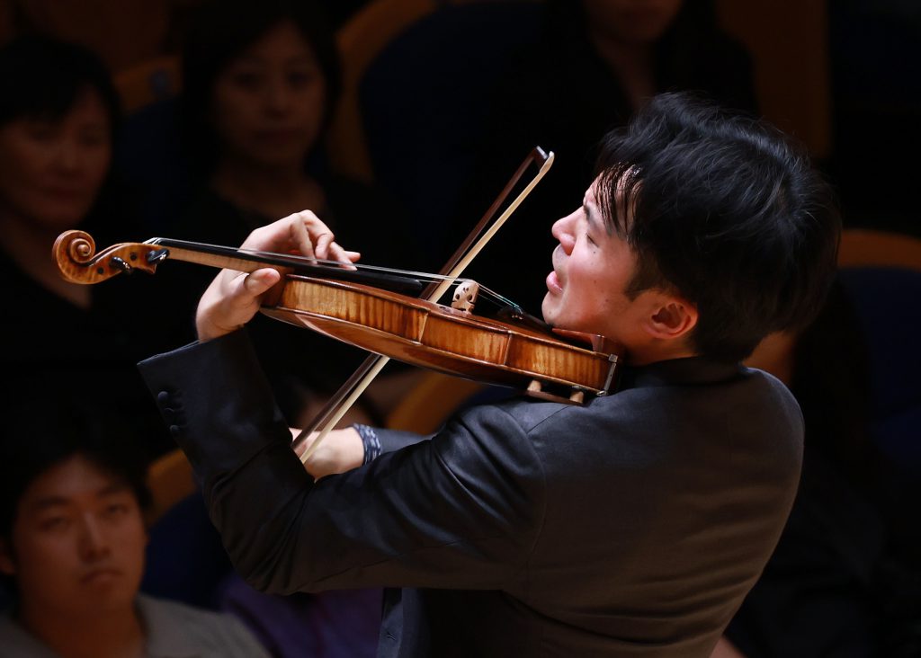 Violinist in a dark suit playing a wooden violin on stage with a dim audience in the background.