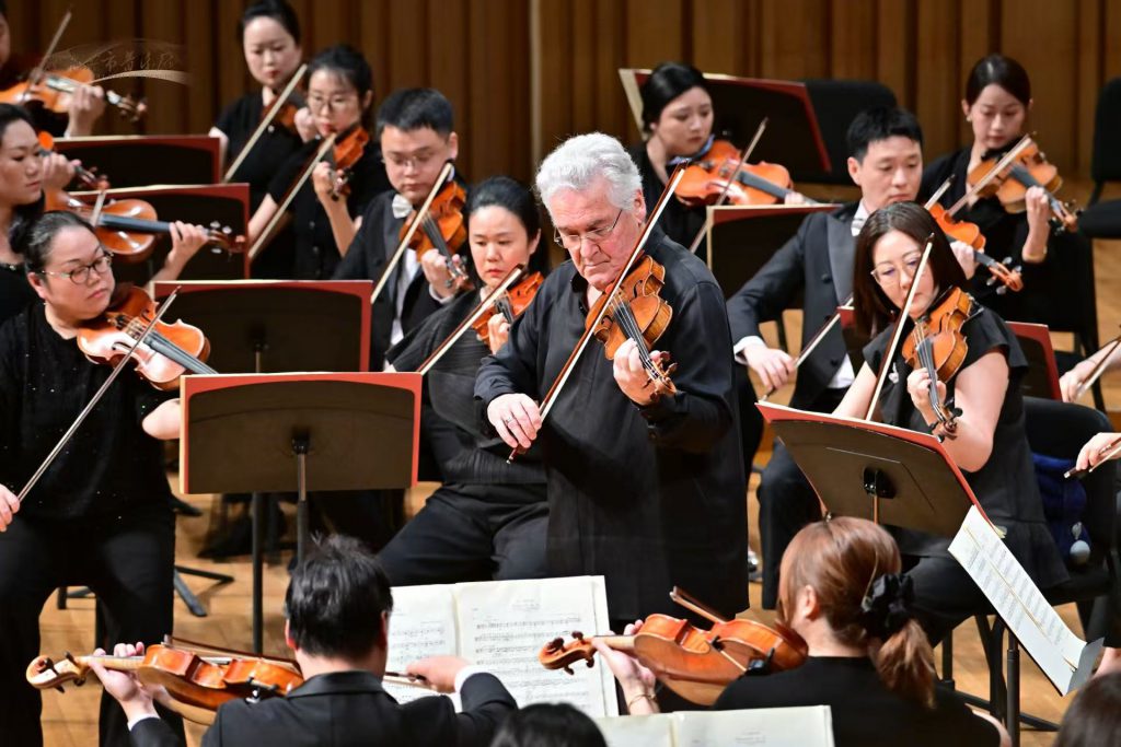 Conductor leads a large string orchestra of violinists in a concert hall.