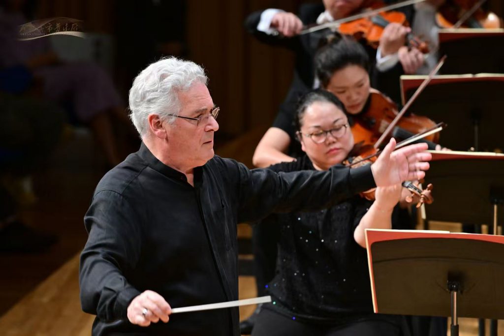Conductor with gray hair guiding an orchestra, baton raised, surrounded by violinists in performance.