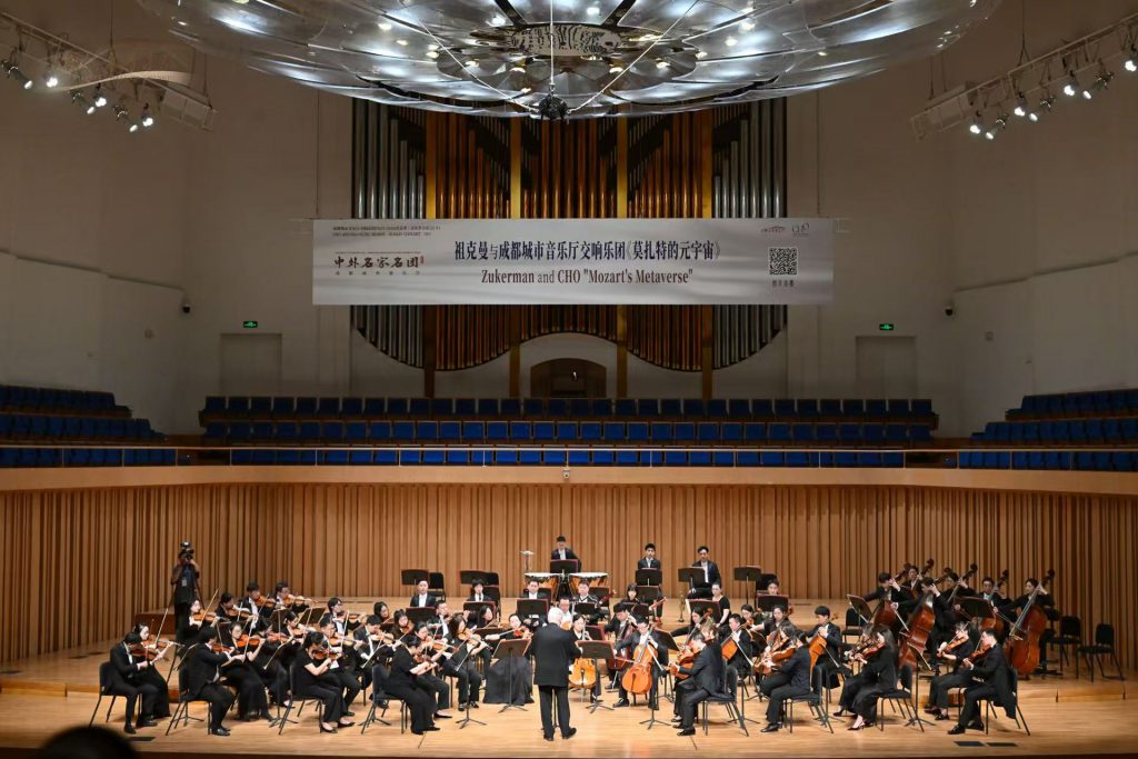Full view of a full orchestra performing on a large concert hall stage, conductor at center, banner overhead reading 'Zukerman and CHO Mozart's Metaverse'.