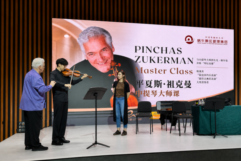 Stage scene with a violinist performing beside an older man and a young woman, against a banner reading Pinchas Zukerman Master Class