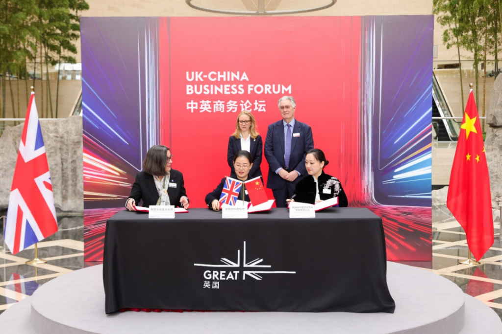 Three signatories at a table signing agreements during a UK–China Business Forum, with UK and Chinese flags on either side and a red backdrop reading UK–China Business Forum.