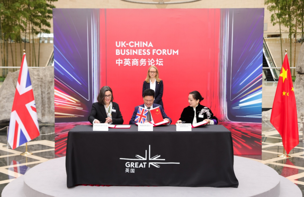 Three delegates sign documents at a signing ceremony for the UK-China Business Forum, with UK and Chinese flags on either side.