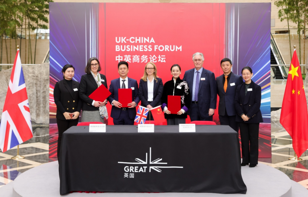 Group of eight professionals standing behind a signing table at a UK–China Business Forum, with UK and China flags on the table and a bold red backdrop.