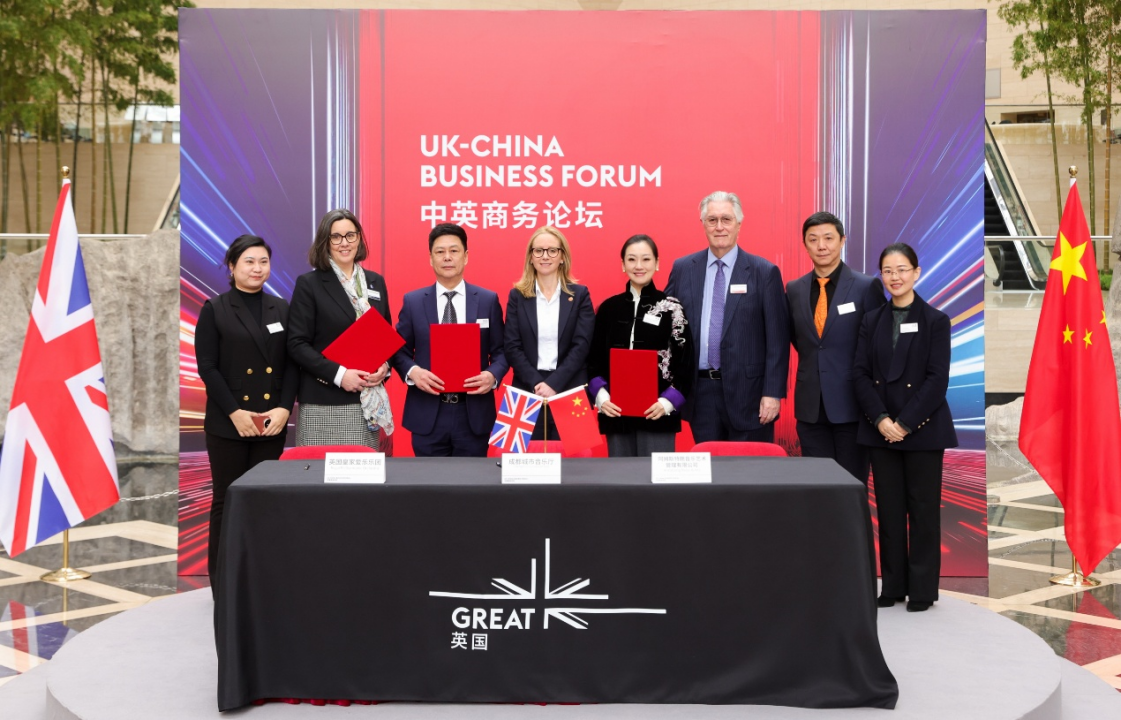 Group of eight professionals standing behind a signing table at a UK–China Business Forum, with UK and China flags on the table and a bold red backdrop.