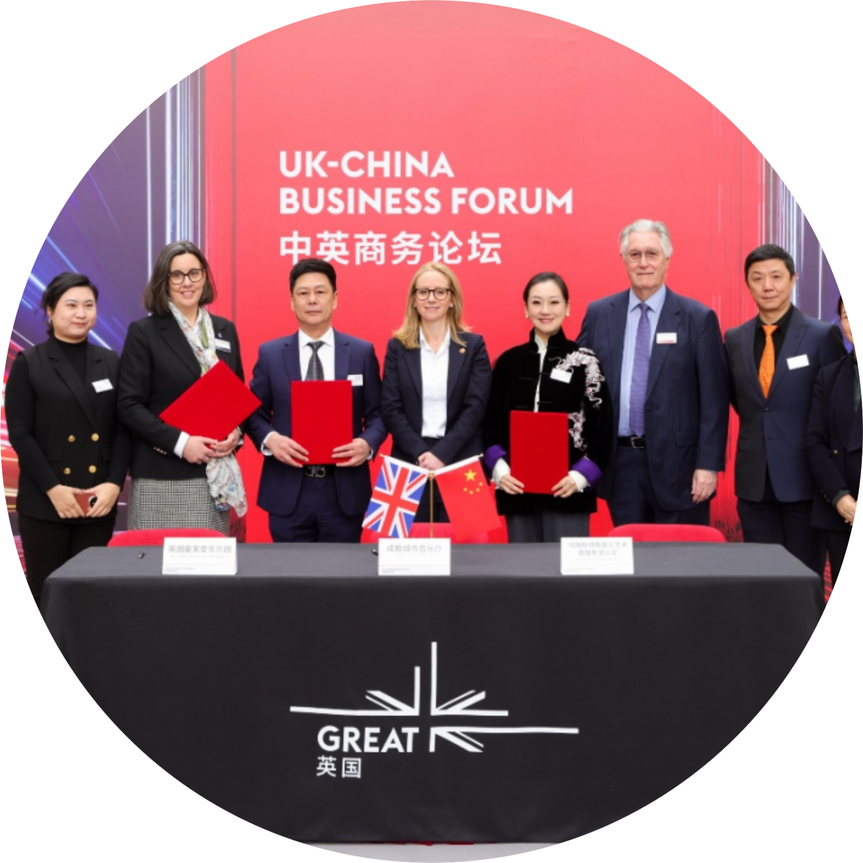 Group of diverse professionals posing with red folders at a UK–China Business Forum event, small flags on the table, red backdrop with Chinese text, circular crop art.