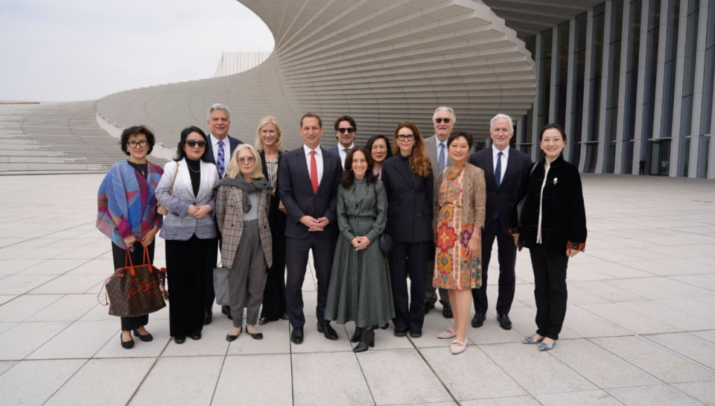 Group of professionals in formal attire posing outdoors on a large tiled plaza with a curved modern building in the background. Group of diverse adults in business clothes standing together for a professional photo outside a futuristic architectural structure. Group portrait of men and women in suits and dresses on a wide plaza, with a spiraling white building behind them. Professional group photo taken outside a contemporary building with a concentric curved design, featuring business people in formal wear. Team of colleagues posing for a photo on a sunny day in front of an architectural complex with white panels.