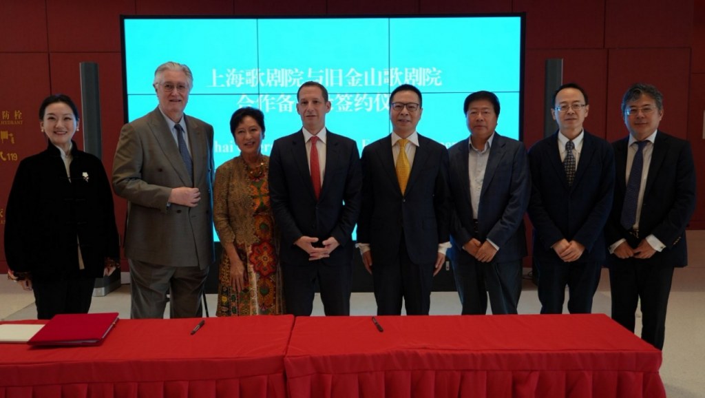 Eight professionals posing for a signing ceremony in front of a blue digital display and red tablecloths, formal attire present.