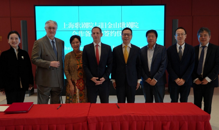 Eight professionals posing for a signing ceremony in front of a blue digital display and red tablecloths, formal attire present.